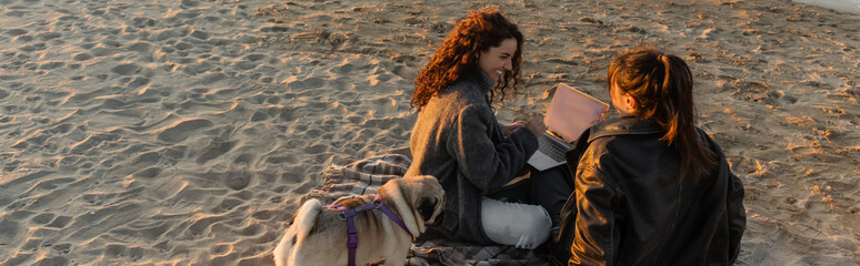 Sticker - Cheerful women using laptop near pug dog on beach during sunset, banner.