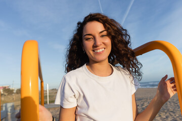 Wall Mural - cheerful young woman with curly hair looking at camera near yellow handrails on beach in Barcelona.