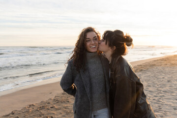 Wall Mural - Young woman kissing cheek of smiling friend on beach in Barcelona.