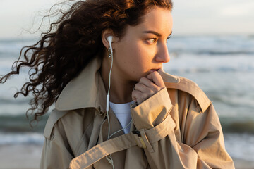 Wall Mural - portrait of curly young woman in beige trench coat and wired earphones listening music near sea in Barcelona.