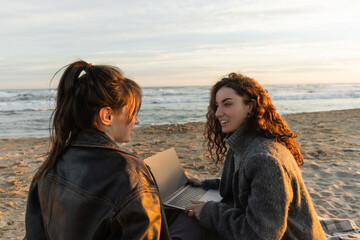 Wall Mural - Smiling woman holding laptop while talking to friend on beach in evening.