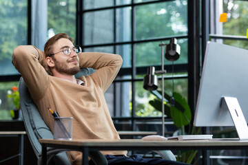 A young male student is taking a break from studying after an online exam. He is sitting in the office at a table with a computer, hands behind his head, leaning back in a chair, smiling.