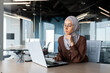 © Liubomir - Successful businesswoman thinking at workplace, muslim woman in hijab using laptop at work, serious arab woman sitting at desk inside office close up.