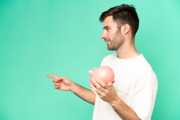 Wall Mural - Young caucasian man holding piggybank isolated on green background pointing to the side to present a product