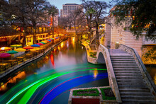 Vibrant Winter Night Landscape Of San Antonio River Walk In Texas With Warm Colors Of Boardwalk, Stone Bridge, Water Reflections, And Light Trails Of River Cruise Tour Boats On The Canal