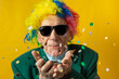 © MarcoDiStefano - Front view of an old man dressed for carnival and blowing confetti with sunglasses, green jacket, colored wig