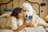 Fototapeta Przestrzenne - Young woman uses smart phone while lying with her cute adorable dog in bed at cozy bedroom in beige tones. Concept of leisure time with pets