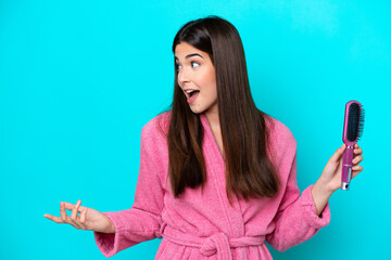 Wall Mural - Young Brazilian woman holding hairbrush isolated on blue background with surprise facial expression