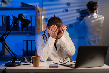 Sleepy doctor with headache working at hospital at the night. Doctor standing in medical clothing near a white wall with board.