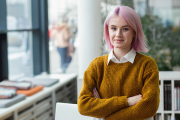 Happy successful modern young woman student, programmer, office worker or entrepreneur stands with crossed arms in a modern office, looking at the camera and smiling friendly