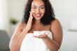 © Prostock-studio - Happy black overweight woman holding jar of organic skin cream, applying moisturiser on face after shower