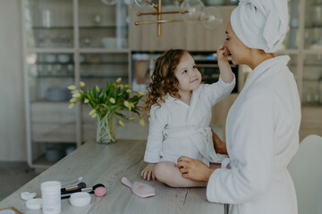 Wall Mural - Pretty curly small girl touches nose of her mother dressed in white soft dressing gown sits on table with cosmetic products going to do makeup for mom pose together against cozy home interior