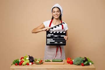 Wall Mural - Young fun housewife housekeeper chef latin woman wear apron toque chefs hat work at table kitchenware hold classic black film making clapperboard isolated on plain beige background. Process cook food.