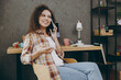 © ViDi Studio - Young cheerful confident smart happy successful employee business woman of African American ethnicity wear casual shirt talk mobile cell phone sit work at office desk with laptop pc computer indoors.