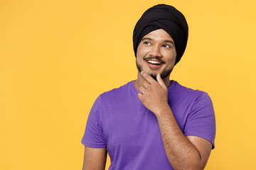 Smiling fun devotee Sikh Indian man ties his traditional turban dastar wear purple t-shirt put hand prop up on chin lost in thought and conjectures isolated on plain yellow background studio portrait