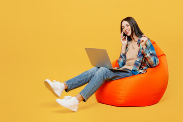 Wall Mural - Full body young woman wear blue shirt beige t-shirt sit in bag chair hold use work on laptop pc computer talk on mobile cell phone isolated on plain yellow background studio. People lifestyle concept.