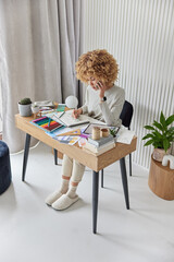 Indoor shot of professional curly haired female interior designer plans design of future apartment asks for advice in colleague via smartphone poses at table makes room drawings works from home