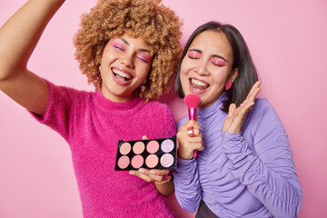 Wall Mural - Happy cheerful multiethnic women foolish around sing song apply eyeshadow palette hold cosmetic brush as if microphone prepare for party isolated over pink background. Beauty treatments concept