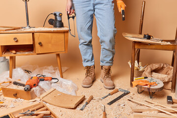 Cropped shot of unrecognizable carpenter in jeans and boots holds instruments poses at messy workshop repairs wooden furniture being professional woodworker surrounded by sawdust. Carpentry concept