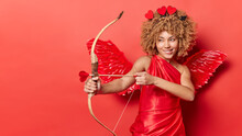 Horizontal Shot Of Cheerful Female Cupid Shoots Arrow Targets On Something Wears Red Dress With Wings Behind Back Looks Mysteriously Somewhere Isolated Over Red Background Copy Space For Your Text