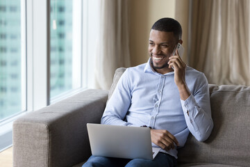 Wall Mural - Cheerful successful Black freelance business man working from home, using digital devices for online communication, making mobile phone call, holding laptop, laughing, looking away with happy smile