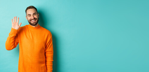 Wall Mural - Photo of friendly young man saying hello, smiling and waiving hand, greeting you, standing in orange sweater over light blue background
