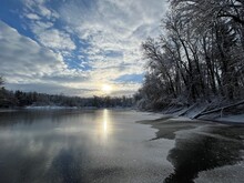 Frozen Pond With Sun In The Centre With Trees Around It And Blue Cloudy Sky Above In Switzerland