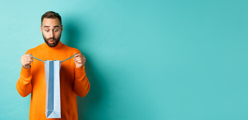 Wall Mural - Surprised man open shopping bag and looking amazed, receiving gift on holiday, standing over turquoise background