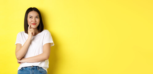 Wall Mural - Beauty and fashion concept. Pensive asian woman thinking, looking thoughtful while pondering something, standing over yellow background