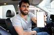 © Alan - young man smiling showing the screen of his phone, sitting on driver's seat, taxi app driver