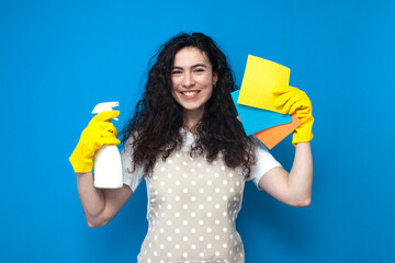 young girl cleaner in uniform holds a spray and rags on a blue background, a woman housekeeper in an apron