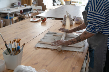 Wall Mural - Woman potter in apron rolls out plate to create piece of art or make handmade utensils. Cropped image of creative girl ceramist working with raw clay learning handicraft standing at table in workshop 