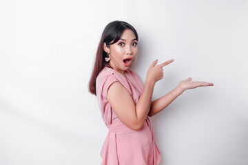 Wall Mural - Shocked Asian woman dressed in pink, pointing at the copy space on beside her, isolated by white background