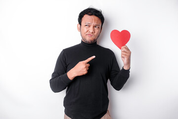 A portrait of an Asian man wearing a black shirt looks so confused while holding a red heart-shaped paper, isolated by a white background