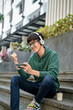 © bongkarn - Cheerful Asian male college student wearing headphones and using his smartphone on street stairs