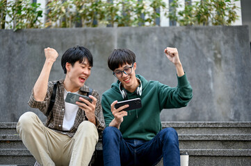 Overjoyed Asian man sits on street stairs watching football match on smartphone with his friend