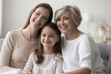 Wall Mural - Cheerful cute child, mother and granny hugging on home couch with love, care, tenderness, enjoying closeness, family meeting, warm relationship, leisure together, laughing, smiling, looking away