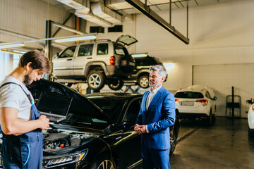 Mature grey hair business male customer with a black car and an auto mechanic looking at a laptop computer in auto-service. Vehicle wiring inspection.