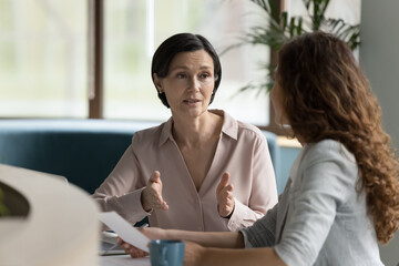 Confident mature business professional woman talking to younger female colleague at office table, speaking, gesturing, teaching, explaining work tasks. Elder mentor training intern