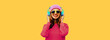 Portrait of happy smiling young woman in wireless headphones listening to music wearing knitted sweater, pink hat on yellow background