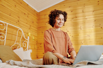 Poster - Younh female freelancer or businesswoman in pajamas sitting on bed in front of laptop in the morning and communicating in messenger