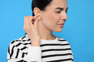 Sticker - Young woman cleaning ear with cotton swab on light blue background, closeup