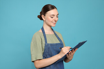 Wall Mural - Beautiful young woman in clean denim apron with clipboard on light blue background