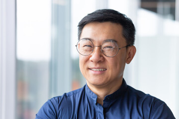 Close-up portrait of smiling asian man inside office, man smiling and looking at camera in glasses and shirt, successful engineer programmer engineer developer in development company at work.