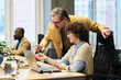 © pressmaster - Two office managers in casualwear discussing data on laptop screen while young woman sitting by desks and mature man bending over her