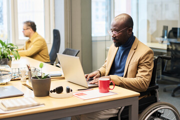 Fototapete - Serious young male broker with disability typing on laptop keypad while sitting by workplace in coworking space and analyzing data