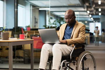 Fototapete - African American businessman with disability sitting on wheelchair with laptop on knees and analyzing online data in openspace office