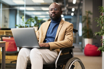 Fototapete - Young serious African American businessman with laptop on his knees sitting in wheelchair in openspace office and looking at camera