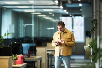 Fototapete - Mature male broker in casual attire using tablet while standing in aisle in openspace office and looking through online financial data