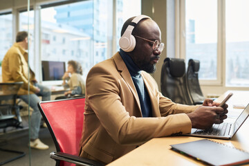 Fototapete - Serious young businessman in headphones choosing new track from playlist in mobile phone while working in front of laptop